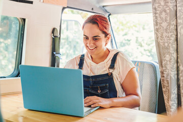 Young woman using her laptop inside her camper van. Van road trip holiday and outdoor summer adventure. Nomad lifestyle concept