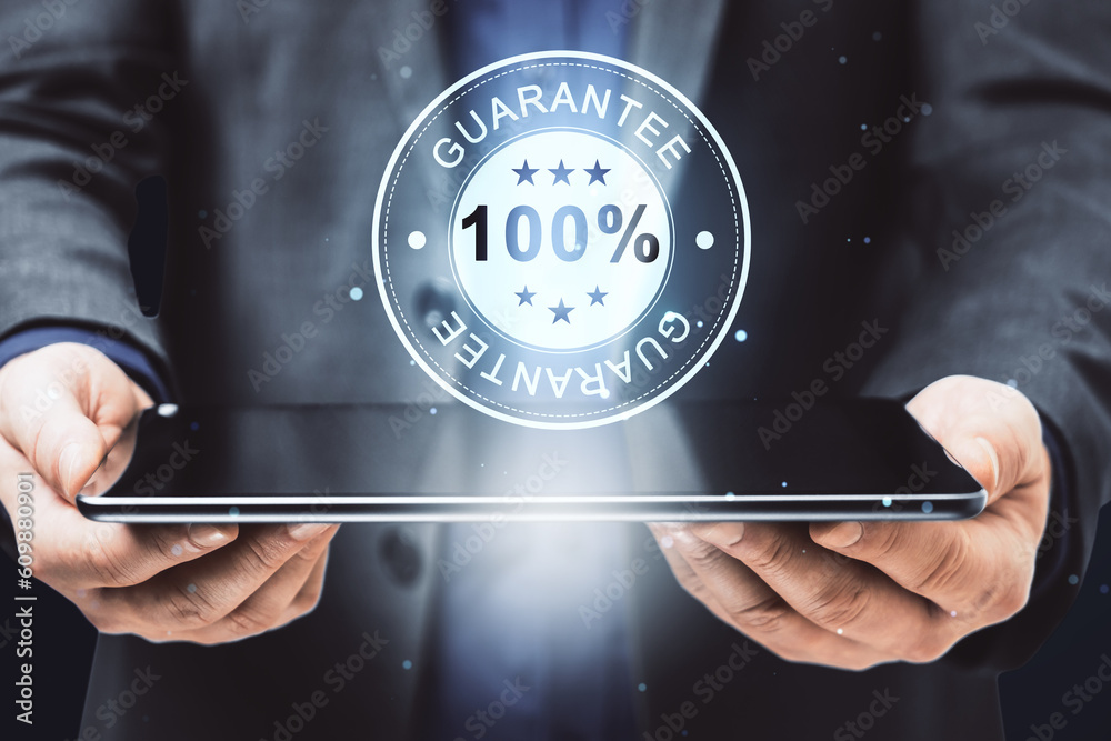 Poster close up of businessman hand holding pad with glowing 100% guarantee stamp hologram on dark backgrou