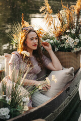 Woman on sunset on picnic by the water in the boat decorated with flowers. Romantic evening portrait of female at her 30s. Relaxation and free time, Floral decorated location