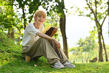 Full length of cheerful young gay man reading book under tree on green grass at sunny beautiful day