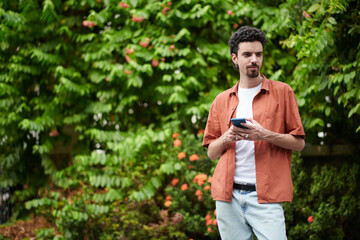 Young man standing outdoors with smartphone in hands and looking away