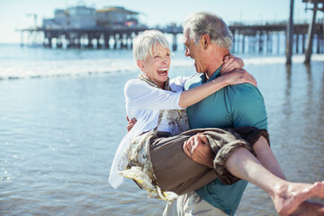 Senior man carrying wife on sunny beach - obrazy, fototapety, plakaty