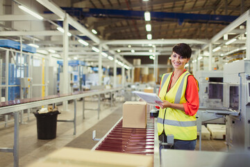 Portrait smiling worker checking cardboard boxes on conveyor belt production line in factory - obrazy, fototapety, plakaty