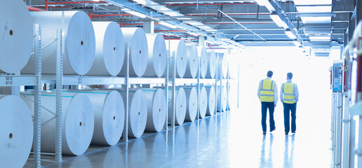Workers in reflective clothing walking along large paper spools in printing plant © KOTO