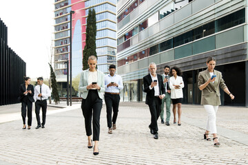 Group of confident business people walking the street using their phones. Diverse team of focused...