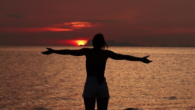 Silhouette of woman looks at sun disk on sky greeting sea sunrise, beginning of new day. Woman enjoying sunrise with her arms outstretched and enjoying beautiful seascape while traveling