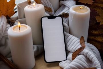 White screen mobile phone app copy space on background of aesthetic atmosphere Autumn leaves spices and candle on knitted white sweater in warm yellow lights. Still life. Raining Outside