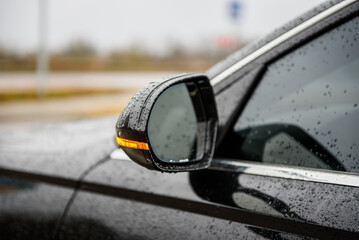 Obraz premium Left rear view mirror in a black car. Side left mirror with turn signal repeater and window of car on street parking and detailing in auto service industry. Road safety while driving. Closeup details.