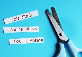Scissors on blue background with paper pieces written YOU SUCK, You're boring, You're Weak - verbal abuse - negative demeaning words abuser uses to gain or maintain power and control over victims - obrazy, fototapety, plakaty