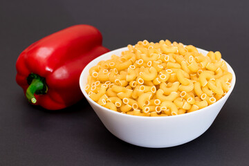 Uncooked Chifferi Rigati Pasta in White Cup and Red Bell Pepper on Black Background. Fat and Unhealthy Food. Scattered Classic Dry Macaroni. Italian Culture and Cuisine. Raw Pasta