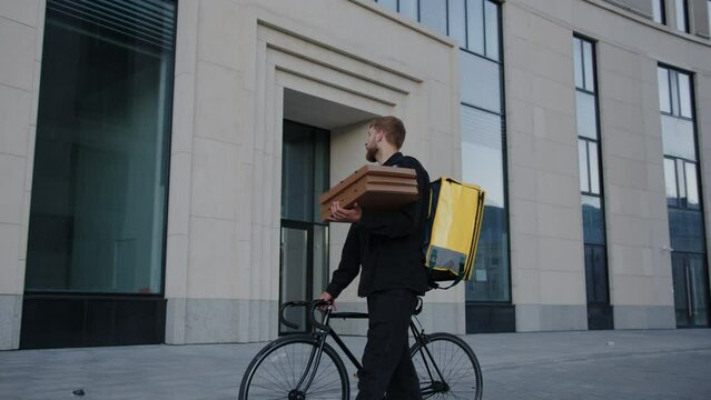 A pizza delivery man walks next to a bicycle through a modern residential complex