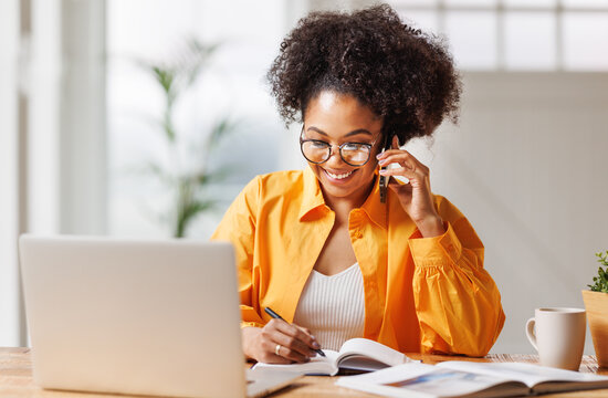 Beautiful Young Smiling Ethnic Woman Making Call Via Smartphone While Working Remotely From Home While Sitting At Desk And Talking To Coworkers .