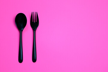 Top view composition of black plastic fork and spoon placed on pink background