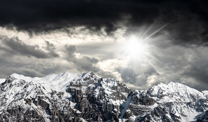 Snow capped mountain peaks in Italian Alps against a dark dramatic sky with sunbeams. Mountain range of Monte Carega (Small Dolomites). Veneto and Trentino Alto Adige, Italy, Europe.