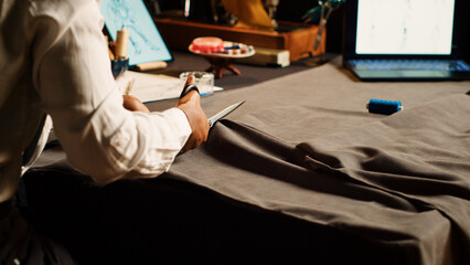African american pupil cutting fabric at workstation, atelier apprentice using scissors to prepare material and sew. Woman working on fashion design collection with industrial tools. Close up.