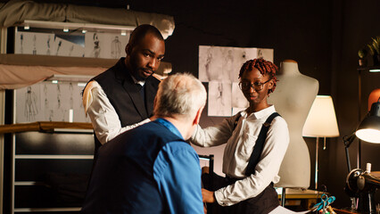 Male client trying on suit blazer in atelier, ensuring right fit and size. Experienced couturier and assistant designing custom made jacket with personal measurements in workshop. Tripod shot.