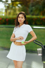 Fototapeta physical activity, young woman with brunette hair standing in stylish outfit with skirt and white polo shirt near cart and holding ball, blurred background, sun-kissed, tennis court in Miami obraz
