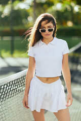 Fototapeta portrait of stylish young woman with brunette long hair standing in white outfit and sunglasses near tennis net, blurred background, wind, tennis court in Miami, iconic city, Florida, sunny day obraz