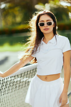 portrait of pretty young woman with brunette long hair standing in white outfit and sunglasses near tennis net, blurred background, tennis court in Miami, Florida, iconic city, sunny day