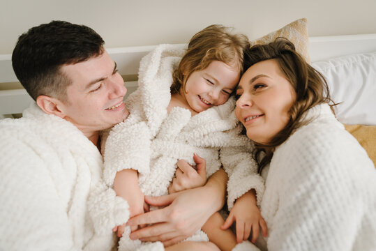 Young mother, father hugs cute little daughter in bathrobes lying on bed, look at each other in hotel room. Happy mom, dad and girl kid face to face and smiling. Top view. Family with child having fun