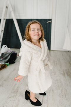 Child Tries On Mother's Shoes Which Are Too Big. Daughter Wearing Mum's Oversize Shoes. Little Model Trying On Clothes At Home. Childhood, Style, Dream Concept. Cute Smiling Blond Girl In Bathrobe.