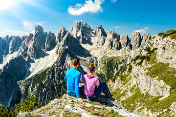 Fototapeta premium Sitting young couple enjoys epic view on Cadini di Misurina mountain range in the morning. Tre Cime, Dolomites, South Tirol, Italy, Europe.
