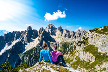 Fototapeta na wymiar Sitting young couple enjoys epic view on Cadini di Misurina mountain range in the morning. Tre Cime, Dolomites, South Tirol, Italy, Europe.