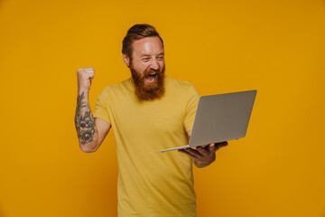 Mid bearded man in yellow shirt screaming and expressing winning gesture holding laptop