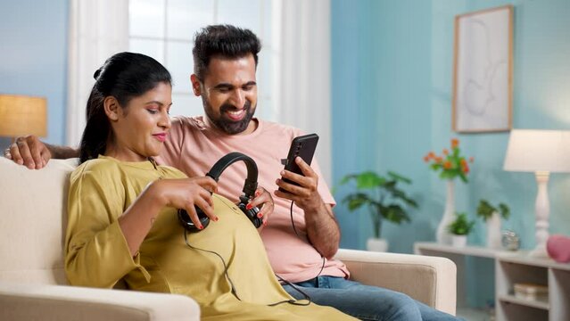 Happy indian husband placing headphones on belly for baby on pregnant wifes belly at home - concept of maternity,