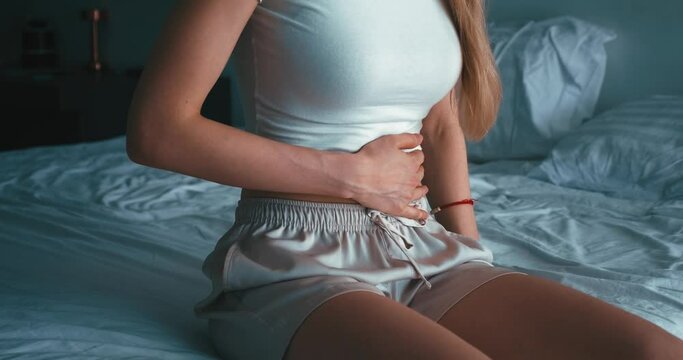 Close up, sick caucasian woman suffer from abdominal pain while sitting in bed, holding stomach, feeling abdominal pain or menstrual pain. Concept of gastritis, diarrhea or painful menstruation