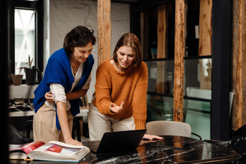 Smiling woman designer and her colleague using laptop while working on interior design project in office