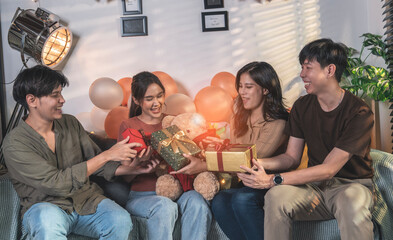 Group of asian friends sitting on sofa, boy friend giving gift box to girl friend celebrating...