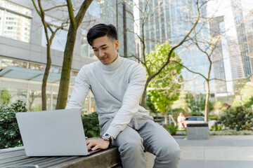 Asian man using laptop, sitting on bench outdoors. Chinese businessman working online, remote work