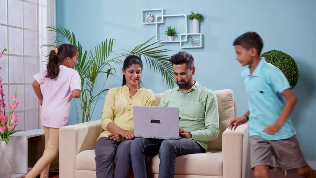 Happy indian kids playing by running around sofa while parents busy on laptop at home - concept of family bonding, mischief and joyful weekend.