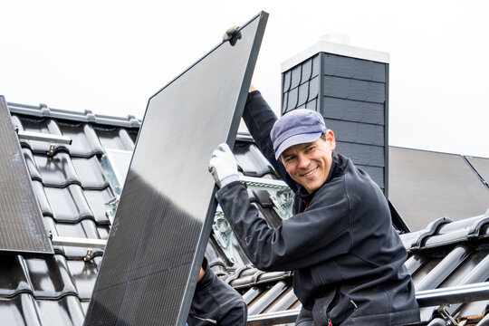 Smiling Tradesman Installing A Solar Panel On A Roof Of A House