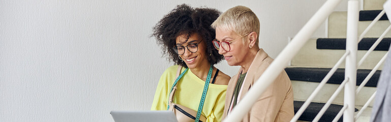 Mature woman and young African-American use laptop stitting on staircase in design studio
