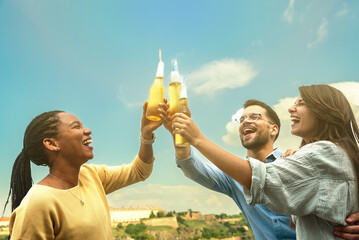 Group of happy young multiracial friends colleagues freelancers in casual clothes drinking cold beer and dancing together while having party on building rooftop at work celebrate end of project