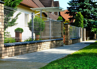 gray metal fence in diminishing perspective. asphalt sidewalk. brick fence piers. decorative green front yard. glass and steel entrance canopy. home ownership concept. lush green pine trees beyond