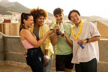 A group of multi-ethnic youths are happily having fun on a one-story rooftop in the open air. The 4 youths are toasting with bottles of beer while wearing party supplies. Young multiracial group. - obrazy, fototapety, plakaty