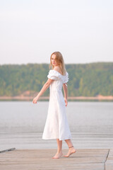Fototapeta na wymiar Portrait of a beautiful girl on the bank of a river. A young woman in a white dress meets a summer sunrise by the water.
