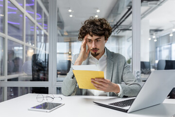 Sad and upset mature businessman received an envelope by mail with a bad news notification letter, boss in business suit working inside office using laptop wearing glasses and beard.