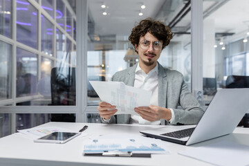 Fototapeta na wymiar Portrait of successful financier at work at workplace inside office, mature man smiling and looking at camera, businessman in shirt working with documents graphs and charts, paperwork using laptop