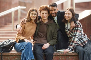 Embracing each other, sitting and smiling. Four young students in casual clothes are together outdoors