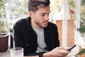 A young handsome man holding a smartphone and looking at the screen, sitting in an outdoor cafe 