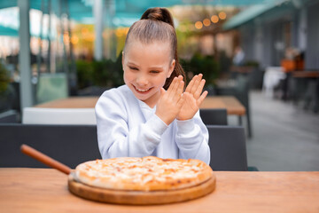 kid girl claps her hands with delight and happiness eating testy italian pizza on wooden board with spatula for cutting
