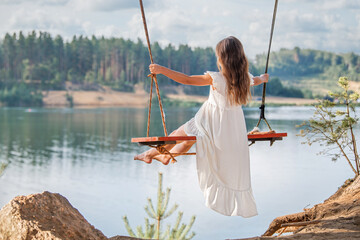 A girl in the forest swinging on a swing. Rope swing on a forest lake. Barefoot girl in a white...