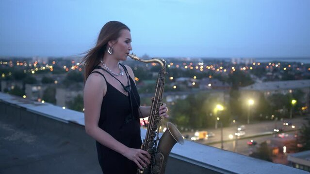 A young woman plays the saxophone while standing on the roof of a building in the city center late at night