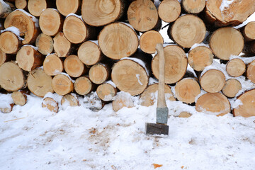 A pile of round firewood, sawn for burning in a furnace, covered with snow, there is an axe next to...