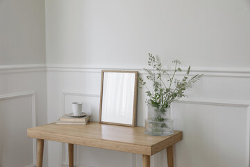 Fototapeta na wymiar Minimal working space, home office. Elegant corner still life. Cup of coffee, books. Empty vertical picture frame mockup. Wooden desk, table. Vase, green grasses and cow parsley. Scandi interior, side
