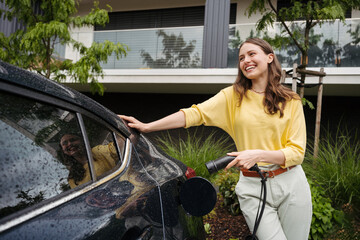 Young woman holding power supply cable from her electric car, prepared for charging it in home,...